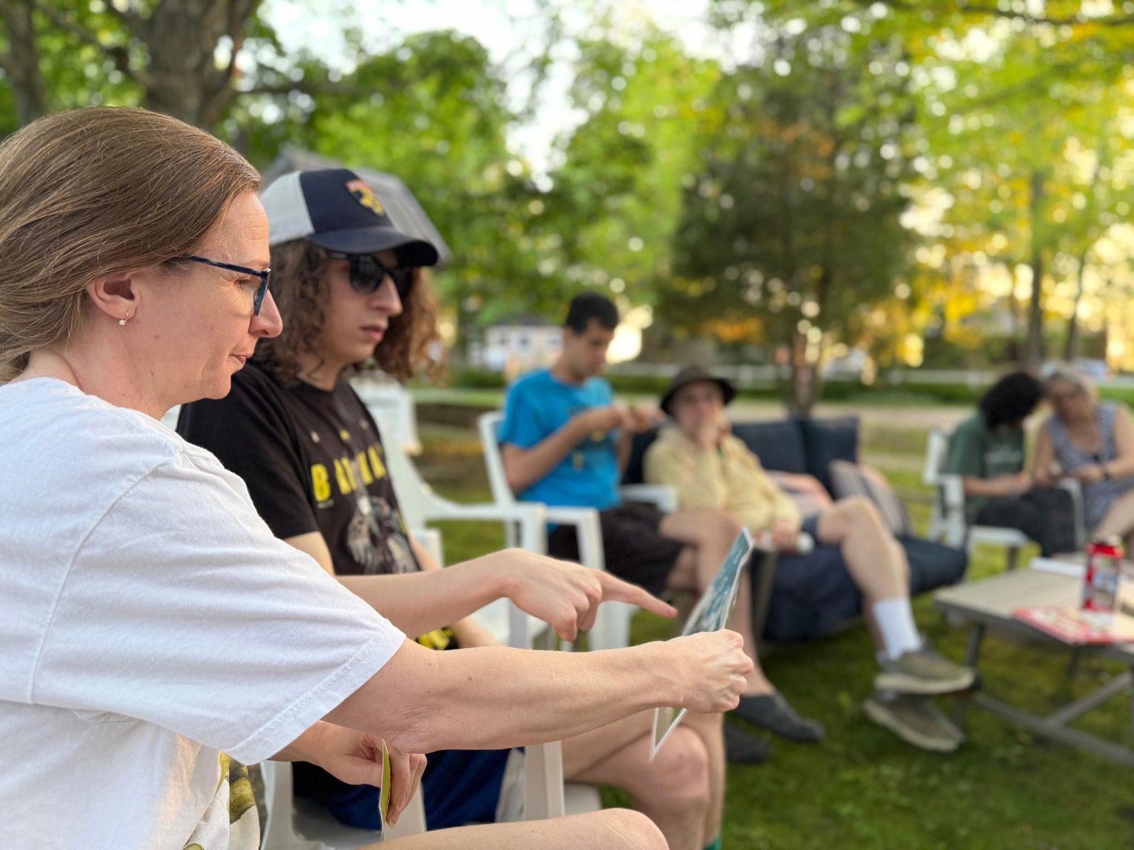 An outdoor meet-up on a summer evening: people sit in a circle of lawn chairs while one of the group points to a card held up to share.